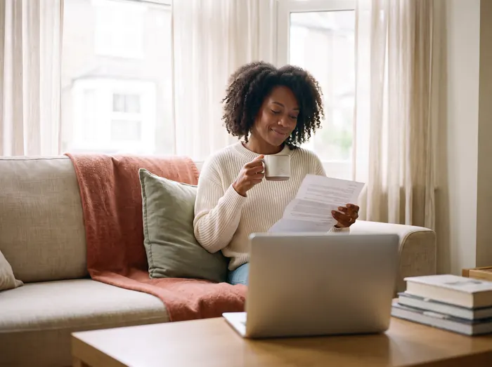 A woman on her sofa with a cup of tea, smiling as she reads a letter